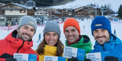 5 junge lachende Leute in bunter Winterkleidung mit Olympia Tickets vor dem Hintergrund eines verschneinten Bergdorfes