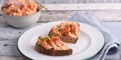 Auf einem hellen Holztisch steht ein weißer Teller mit grünem Rand. Darauf liegen zwei Scheiben dunkles Brot, die großzügig mit einer rosa-orangenen Creme bestrichen sind. Die Creme wirkt locker und ist mit etwas Paprikapulver und kleinen grünen Kräuterstückchen garniert. Im Hintergrund steht eine Schüssel mit derselben Creme. Daneben liegt eine gefaltete, hellblaue Stoffserviette. Die Szene wirkt wie eine appetitliche Präsentation eines Brotaufstrichs.