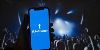„Eine Person hält ein Smartphone mit der Ticketmaster-App auf dem Bildschirm. Im Hintergrund ist unscharf ein Konzert mit Bühne, Lichtstrahlen und jubelndem Publikum zu sehen.