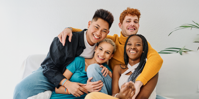 Vier junge Erwachsene sitzen eng zusammen auf einem Sofa und lächeln in die Kamera. Die Gruppe wirkt vertraut und entspannt; im Hintergrund ist eine helle Wand mit einer Pflanze zu sehen.