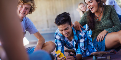 Mehrere Jugendliche sitzen zusammen im Freien, lachen und schauen auf ein Smartphone. Einer hält das Handy, während die anderen über seine Schulter mitsehen.