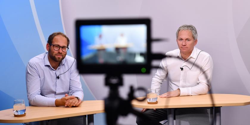 Zwei der Panelteilnehmer, die vor der Kamera an einem Tisch sitzen.