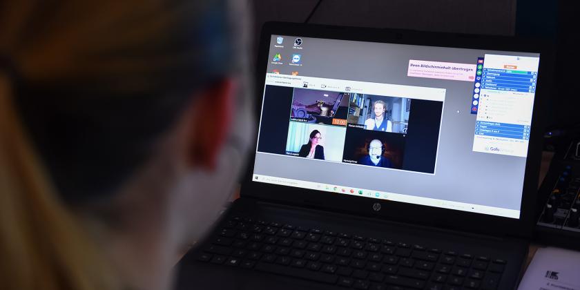 Eine Dame, die auf einen Computerbildschirm schaut. Auf diesem ist ein Videocall zu sehen.