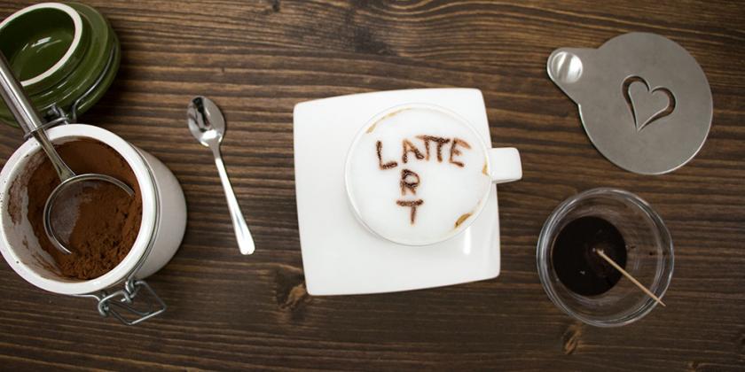 Latte Art: Kaffeetasse mit Milchschaum - rundherum Latte-Art-Utensilien, Becher mit Schokosauce, Herzschablone, Zahnstocher in Schokosauce getaucht zum Schreiben auf Milchschaum