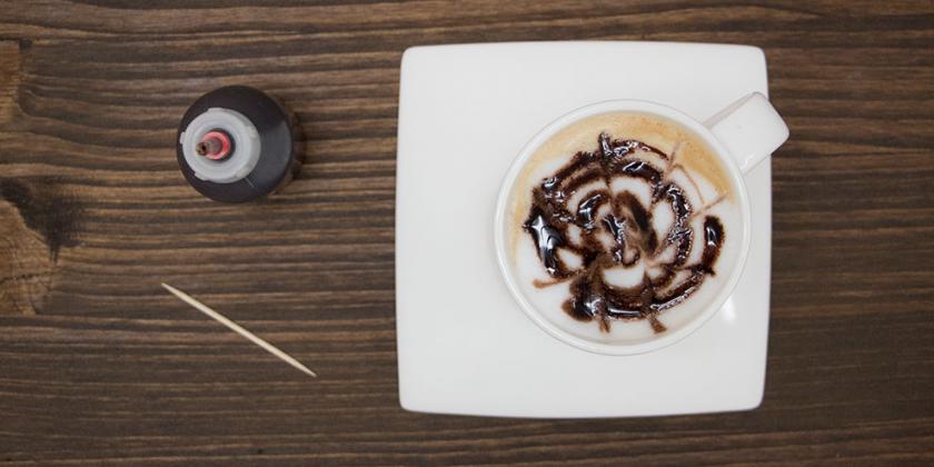Latte Art: Kaffeetasse mit Milchschaum, obenauf Schokosirup als Verzierung, kl. Fläschchen mit Schokosirup steht am Tisch neben der Tasse