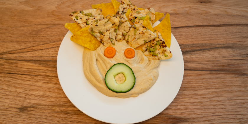 Hummus auf Teller mit Gemüsegesicht und Tortillachips-Frisur
