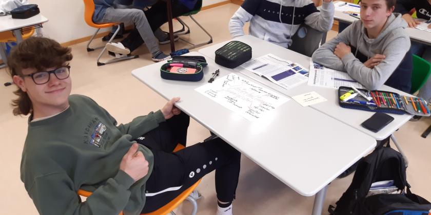 Schüler an Schreibtischen mit Unterlagen und KONSUMENT