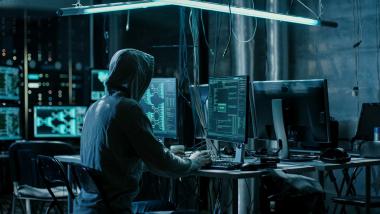 Person with dark hood makes inputs in front of a computer in a dark room with many screens