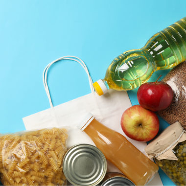 Lebensmittel wie Nudeln, Öl, Apfelsaft, Konserven und Äpfel liegen neben einer weißen Papiertüte auf blauem Hintergrund.