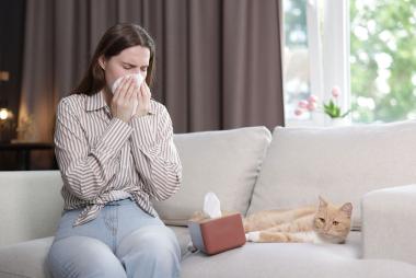 Junge Frau sitzt am Sofa mit einer Katze und putzt sich die Nase.