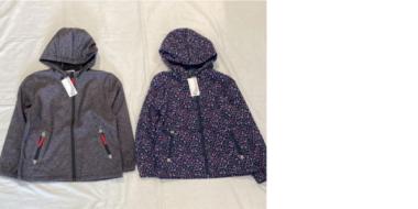 2 Jacken, die nebeneinander auf einem hellen Untergrund liegen.  Linke Jacke: Dunkelgraue Softshelljacke mit Kapuze. Sie hat zwei Reißverschlusstaschen mit roten Reißverschlüssen und eine weiße Etikette hängt am Kragen.  Rechte Jacke: Dunkelblaue oder lilafarbene Jacke mit Kapuze, bedruckt mit einem kleinen, bunten Punkt- oder Blümchenmuster. Auch sie hat zwei Reißverschlusstaschen und ein Etikett am Kragen.