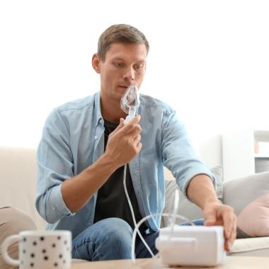 Ein Mann sitzt auf einem Sofa, vor sich steht ein Inhalationsgerät, das er bedient und das zugehörige Mundstück vor seinen Mund hält. 