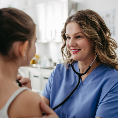 Eine Ärztin in blauer Kleidung mit Stethoskop untersucht ein junges Mädchen in einem grauen Trägertop.