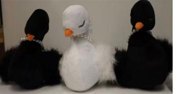 Drei Stofftiere in Form von Schwänen sitzen nebeneinander auf einer ebenen Fläche. Der mittlere Schwan ist weiß, die beiden äußeren sind schwarz. Alle drei haben orangefarbene Schnäbel und geschlossene Augen mit langen, geschwungenen Wimpern, was ihnen einen friedlichen Ausdruck verleiht. Um ihre Hälse tragen sie Perlenketten. Die Schwäne sind weich und flauschig gestaltet, mit einem stilisierten, eleganten Design.