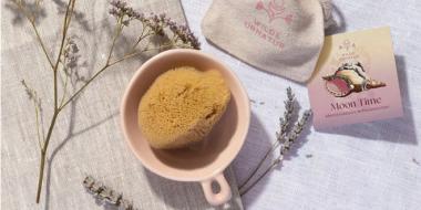 Ein liebevoll arrangiertes Stillleben zeigt natürliche Selbstfürsorgeprodukte: Ein Meerschwamm in einer rosa Tasse, ein Stoffbeutel mit floraler Grafik („WILDE URNATUR“) und die Kräutermischung „Moon Time“ zur Menstruationsunterstützung. Getrockneter Lavendel rundet die ruhige, achtsame Atmosphäre ab.