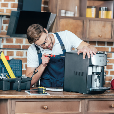 Ein Mann in Arbeitskleidung repariert mit einem Schraubenzieher eine Kaffeemaschine auf einer Werkbank. Daneben liegen Werkzeug, Maßband und ein Werkzeugkoffer; im Hintergrund ist eine Backsteinwand zu sehen.