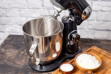 Küchemaschine mit Schwenkarm auf einer Holzarbeitsfläche, daneben ein Brett mit Holzschüsseln und Mehl sowie Salz, im Hintergrund eine weiße Wand.