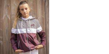 Blondes Mädchen steht mit leichter Reitjacke vor eine Holzwand