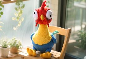 Auf dem Bild ist eine bunte, comicartige Hühnerfigur zu sehen, die auf einem kleinen Holzstuhl sitzt. Das Huhn hat einen roten Kamm, große hervorstehende Augen und einen gelben Schnabel. Der Körper ist blau und gelb gefärbt, die Füße sind gelb. Die Szene wirkt freundlich und verspielt. Im Hintergrund erkennt man ein helles Zimmer mit großen Fenstern und grünen Pflanzen, durch die warmes Tageslicht einfällt.