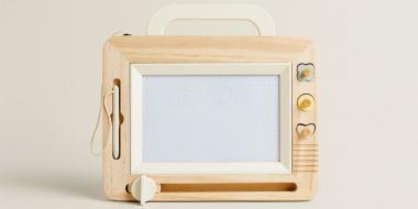 Eine Tafel mit hellem Holzrahmen steht vor einem neutralen Hintergrund. Es erinnert an einen alten Fernseher oder ein Radio, mit Tragegriff oben, seitlichen Drehknöpfen und einem rechteckigen, hellen Bildschirm, der als Zeichenfläche dient..