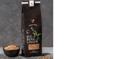 Auf dem Bild steht eine matte, dunkelbraune Packung „Bio-Berglinsen“ der Marke Wiener Gusto auf einem grauen Untergrund. Auf der Vorderseite sind ein grüner Pflanzenzweig, ein Sichtfenster mit den Linsen sowie mehrere Logos abgebildet. Links daneben steht eine kleine schwarze Schale, gefüllt mit hellbraunen Linsen. Einige Linsen sind locker auf der Fläche verstreut.