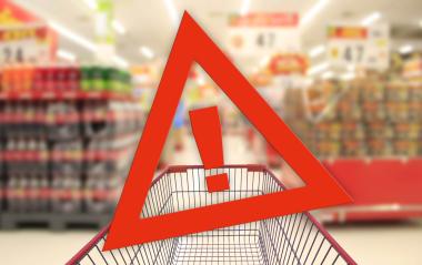 Ein Einkaufswagen steht in einem Supermarkt, der Hintergrund ist unscharf. Darüber liegt ein großes rotes Warnsymbol in Dreiecksform mit einem Ausrufezeichen in der Mitte.