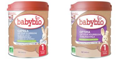Auf dem Bild sind zwei Dosen Babybio-Säuglingsmilch zu sehen. Links steht eine Packung „Caprea“ aus Ziegenmilch, rechts eine Packung „Optima“. Beide Behälter sind zylindrisch, haben rote Deckel und ein naturfarbenes Etikett. Jede Dose zeigt das Babybio‑Logo, Produktinformationen in mehreren Sprachen sowie ein kleines Stofftier als Illustration.
