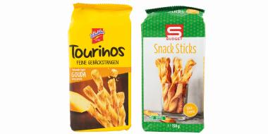 Die Abbildung zeigt zwei aufrecht stehende Snackverpackungen mit Gebäckstangen vor weißem Hintergrund. Links befindet sich eine leuchtend gelbe Packung mit der Aufschrift „Tourinos – Feine Gebäckstangen“ sowie einem Hinweis auf Gouda‑Geschmack. Das Produktbild zeigt mehrere knusprige Stangen. Rechts steht eine grüne Verpackung mit Punktmuster und rotem Logo; darauf sind „Snack Sticks“ sowie ein Glas mit gedrehten Gebäckstangen abgebildet. Beide Produkte wirken ähnlich positioniert und vergleichbar groß.