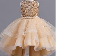 Festliches, hellgoldenes Kleid auf einem Kleiderständer oder einer Puppe. Das Oberteil besteht aus dicht angeordneten, glitzernden Pailletten in Goldtönen. Der Rock ist mehrlagig und voluminös, aus transparentem Tüll mit feinen, funkelnden Punkten. Die obere Lage des Rocks ist asymmetrisch geschnitten, kürzer vorne und länger hinten. In der Taille befindet sich ein schmaler, dekorativer Gürtel mit kleiner Schleife.