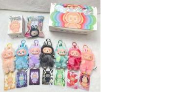 Verschiedene bunte Anhänger und kleine Figuren zu sehen, die auf einer hellen Oberfläche angeordnet sind. Oben liegen zwei Verpackungen mit pastellfarbenem Design und verspielten Motiven. Darunter befinden sich mehrere Schlüsselanhänger in Form von kleinen, plüschigen Figuren mit unterschiedlichen Farben wie Blau, Rosa, Grün und Schwarz. Jede Figur hat ein eigenes Gesicht und Design, oft mit großen Augen und süßen Details. Unten sind zusätzlich Karten oder Verpackungen mit passenden Illustrationen ausgelegt