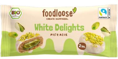 Auf der Verpackung sind 2 Snacks der Marke „foodloose“ mit dem Namen „White Delights Pistazie“ zu sehen. Die 2 runden Snacks sind von weißer Schokolade überzogen und mit Pistazienstückchen bestreut. Ein aufgeschnittener Riegel zeigt die grüne Füllung im Inneren. Zudem sind Bio- und Fairtrade-Siegel abgebildet.