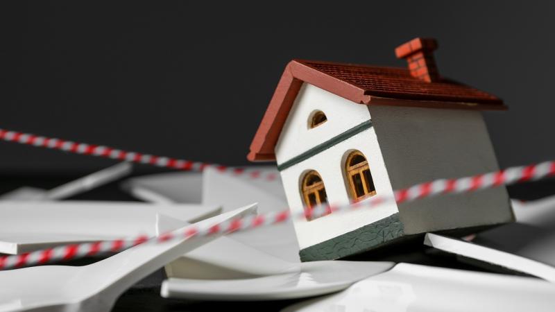 Modellhaftes Haus, das auf zerbrochenen Scherben steht. Eingefriedet mit Absperrband.