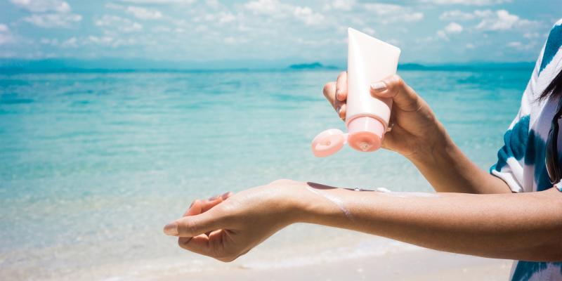 Frau hält Sonnencreme-Tube in der Hand und trägt Creme auf ihre linke Unterarminnenseite auf, dahinter türkisblaues Meer