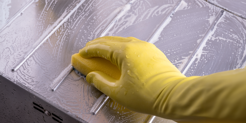 Hand in gelbem Gummihandschuh und gelbem Schwamm wischt über Edelstahlabtropffläche einer Abwasch