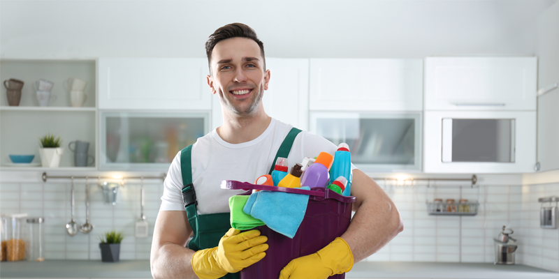 Junger mann in weißem Leiberl und Latzhose posiert in gelben Gummihandschuhen mit Reinigungsmaterialien gefülltem Kübel