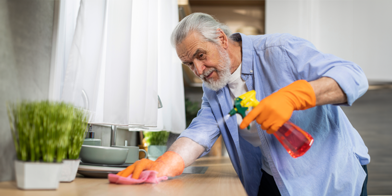 Älterer grauhaariger Mann mit Vollbart in blauem offenen Hemd und Gummihandschuhen sprüht mit linker Hand Reiniger auf Küchenarbeitsplatte, die rechte mit Wischtuch auf Arbeitsplatte abgestützt
