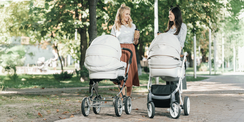 Zwei Frauen schieben je einen hellen Kombikinderwagen in einem Park.
