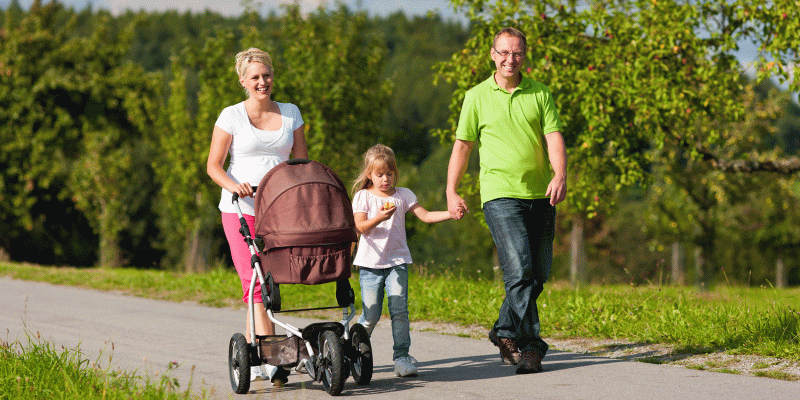 Eine fröhliche Familie spaziert bei sonnigem Wetter auf einem Weg durch eine grüne Landschaft. Die Mutter schiebt einen Kinderwagen, die Tochter geht zwischen den Eltern und hält die Hand des Vaters. Alle wirken entspannt und genießen die gemeinsame Zeit im Freien.