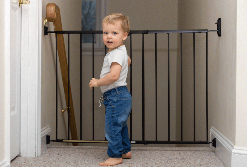 Blonder kleiner Bub in blauer Hose und weißem Leiberl steht vor einer Treppe, die mit einem Treppenschutzgitter gesichert für ihn nicht zugänglich ist