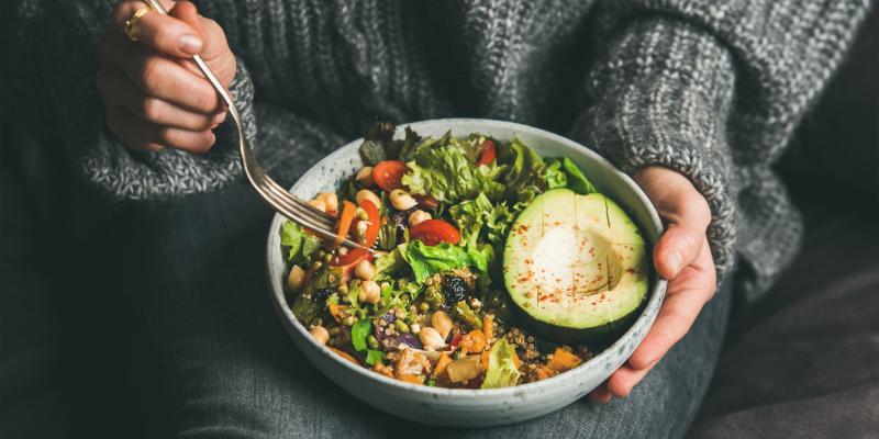 Eine Schüssel mit verschiedenen Gemüsesorten zubereitet zu einer Bowl, eine Frau in grauem Pullover fährt mit einer Gabel in die Schüssel.