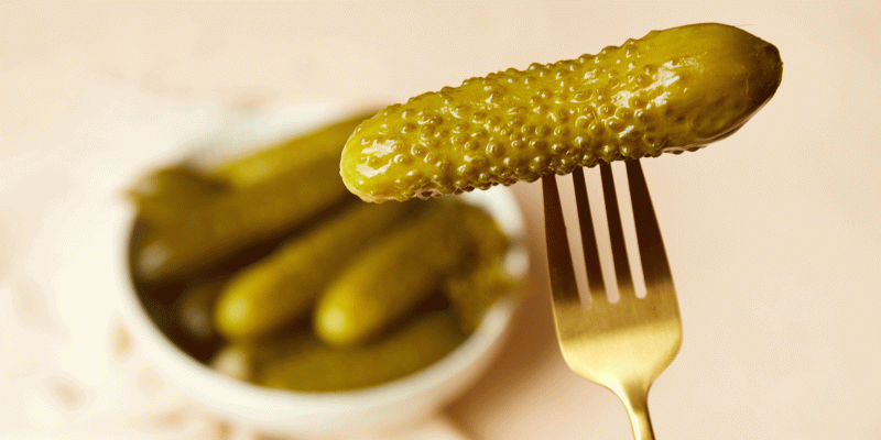 Ein auf einer goldfarbenen Gabel aufgespießtes Essiggurkerl, im verschwommenem Hintergrund eine weiße Schale mit Essiggurkerln