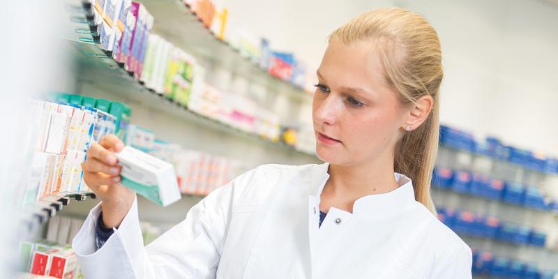 Apothekerin nimmt Packung von Glasregal, im Hintergrund viele Regale Medikamente