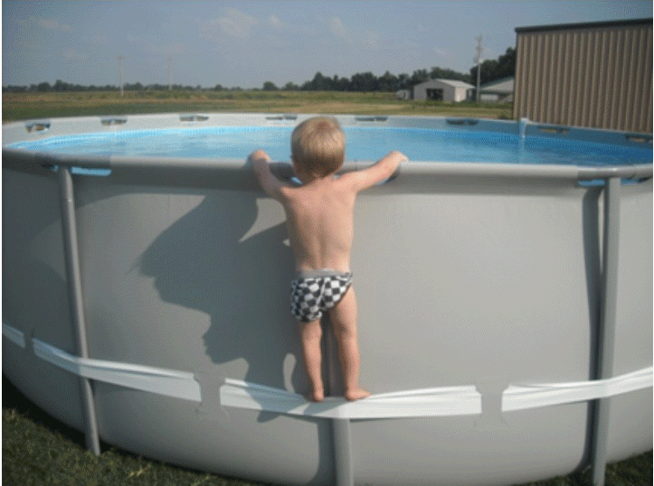 Ein Kind steht barfuß auf dem Kompressionsriemen eines großen, runden Pools und hält sich am oberen Rand fest. Es trägt karierte Badesachen und blickt neugierig ins Wasser. Der Pool steht draußen auf einer Wiese, im Hintergrund sind Felder und Gebäude zu sehen.