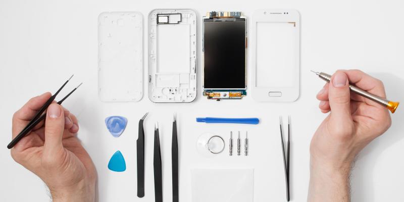 Ein Handy liegt in die Einzelteile zerlegt auf einem Tisch. Davor liegen diverse Spezialwerkzeuge und man sieht die Hände einer Person, die eine Pinzette und eine Schraubenzieher in der Hand hält