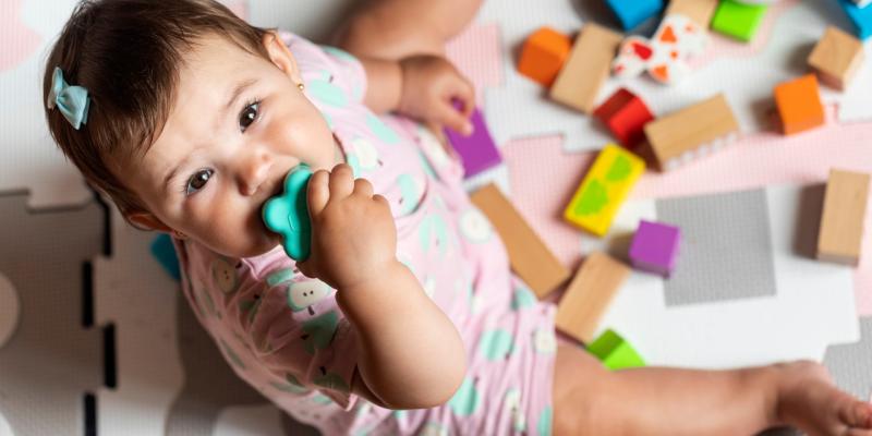 Ein Kleinkind sitzt auf einem Teppich, rund um das Kind herum liegt Spielzeug, eines davon hat es im Mund. 