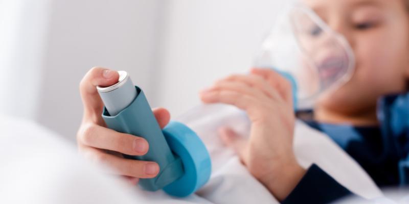 Ein Kind hat einen Asthmaspray mit Aufsatz in der Hand und hält es an seinen Mund. 