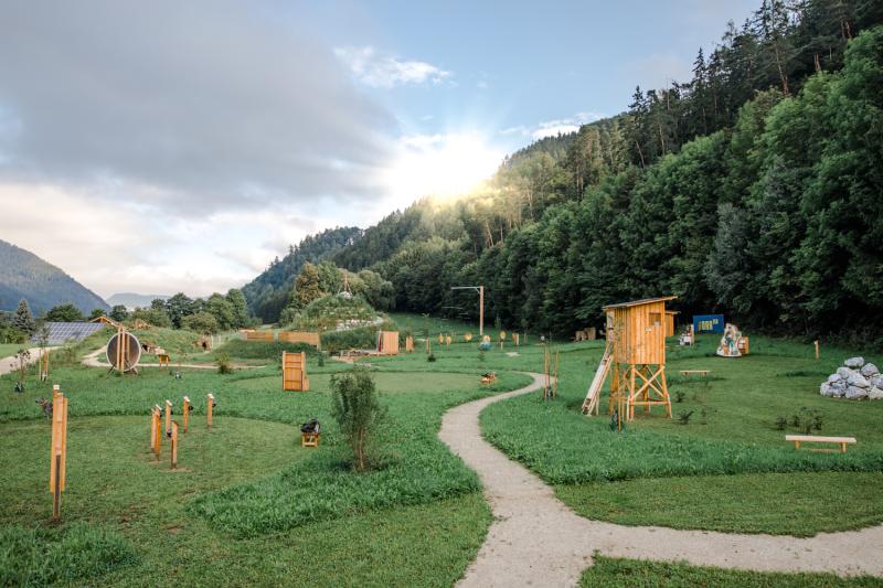 Ebenes, barrierefreies Areal mit Wegen durch Wiesen mit Stationen der Erlebniswelten Steiermark. Die Sonne blitzt zwischen Wolken und bewaldetem Horizont durch.