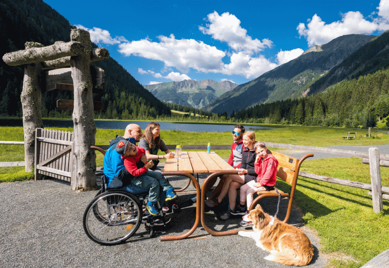 Familie mit Mann im Rollstuhl, auf ihm ein Kind, sitzt mit Hund an einem Tisch einer gepflegten Anlage des barrierefreien Themenweges in einem schönen Alpental.