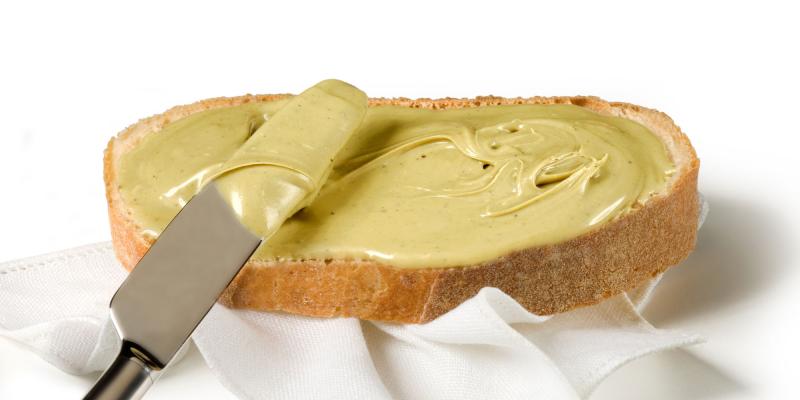 Scheibe Weißbrot mit Pistaziencreme bestrichen auf weißer Serviette und weißem Untergrund. Über dem Brot ist ein halb mit Pistaziencreme bedecktes Messer zu sehen.