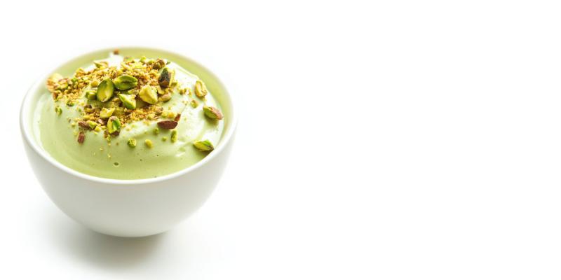 Eine weiße Schale, gefüllt mit hellgrüner Pistaziencreme oder Mousse. Obenauf sind grob gehackte Pistazien verteilt. Die Schale steht links im Bild, der Hintergrund ist komplett weiß.